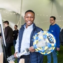 A person smiling broadly holds a large, colorful geometric object with clear panels in an event tent. Other attendees are visible in the background, engaged in activities and discussions. 帐篷用串灯装饰着.